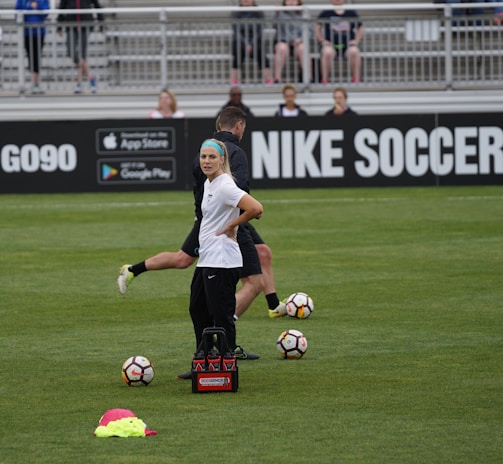 A person in sports attire stands on a soccer field, surrounded by several soccer balls and a carrier with bottles. The field is bordered by advertisements for Nike Soccer and app stores. Several people sit in bleachers in the background.