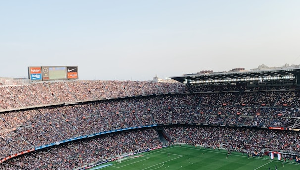 An expansive soccer stadium filled with thousands of spectators. The green field in the center features players and markings, surrounded by vibrant stands and advertising boards on the upper sides of the stadium. The audience is actively watching the game.